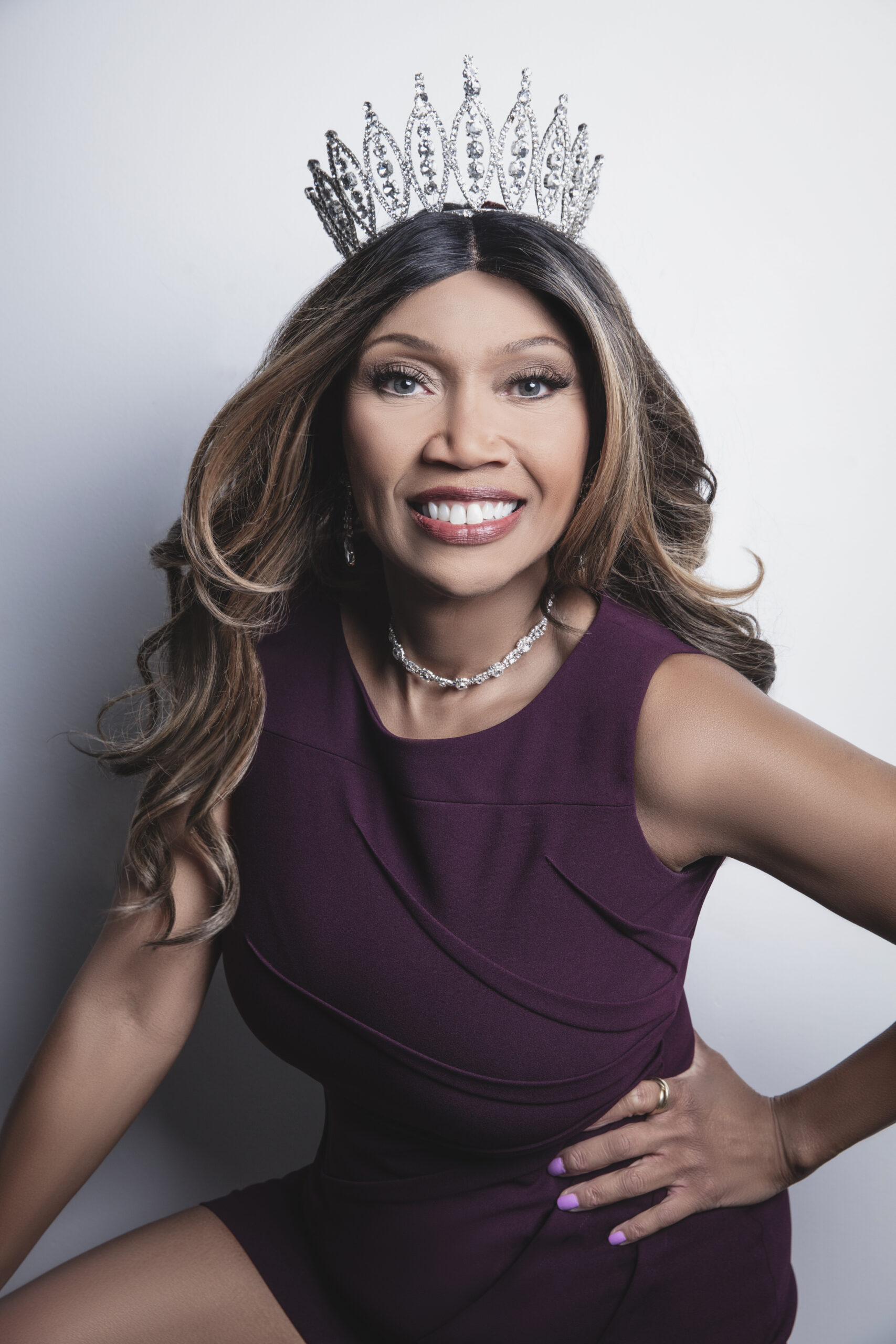 A smiling woman wearing a jeweled crown and a purple dress poses confidently against a light gray background, exuding elegance and joy.
